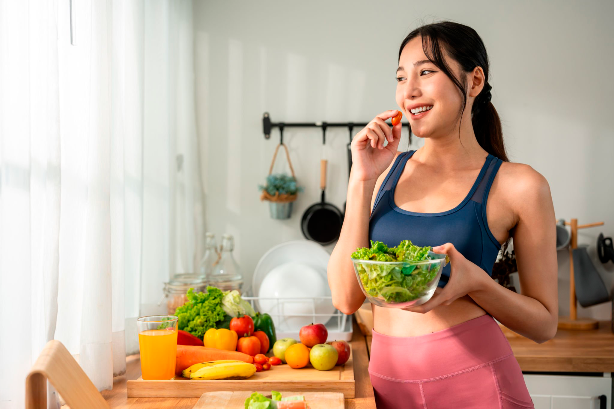 Mujer joven en ropa deportiva comiendo una ensalada saludable en su cocina, rodeada de frutas y verduras, promoviendo una alimentación balanceada para prevenir el deterioro cognitivo