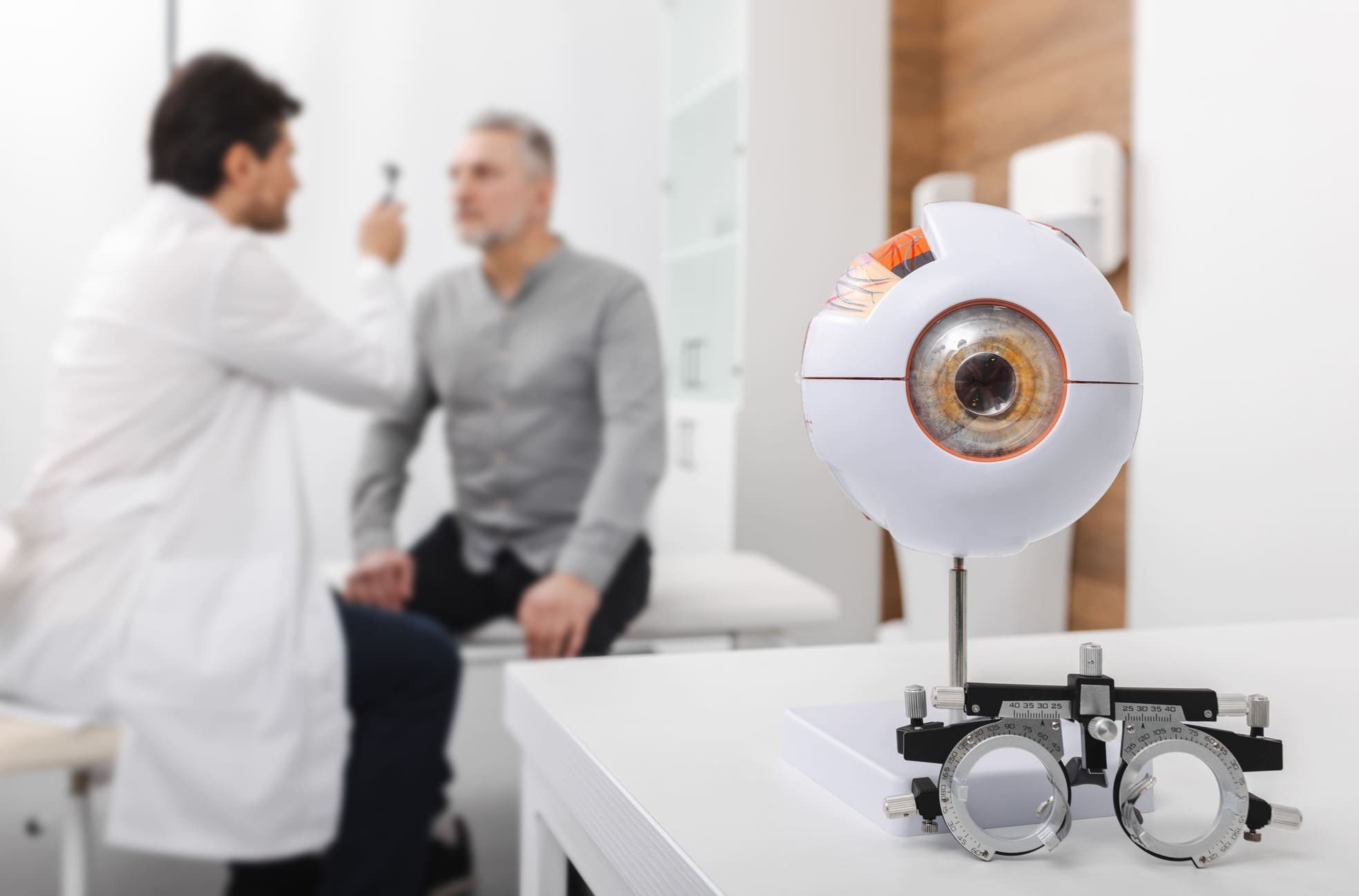 Hombre sentado en una consulta médica mientras su doctor revisa su visión y en primer plano se ve un aparato de medición de dioptrías y una maqueta de un ojo.